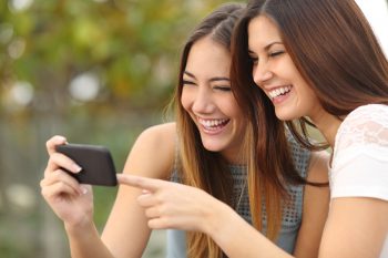 Two funny women friends laughing and sharing social media videos in a smart phone outdoors. Courtesy
